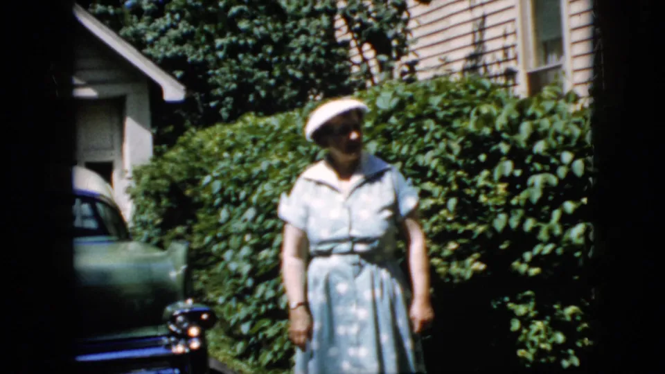 At the end of the 1969 California clip, the woman remains in the driveway, partly shadowed by the 8mm frame edge, with shrubs, siding, and a partial car still visible.