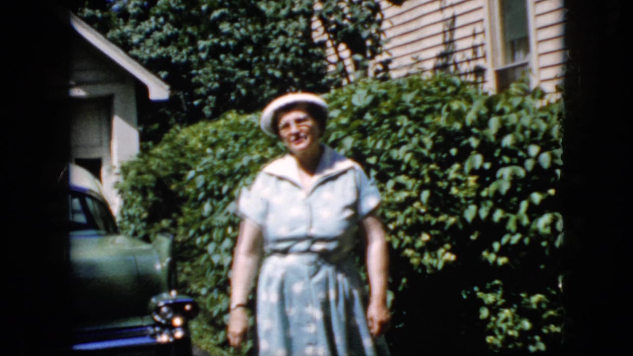 Adult woman in a white hat and light blue polka-dot dress faces the camera in a sunlit California driveway, with a green car, hedges, and house siding behind her.