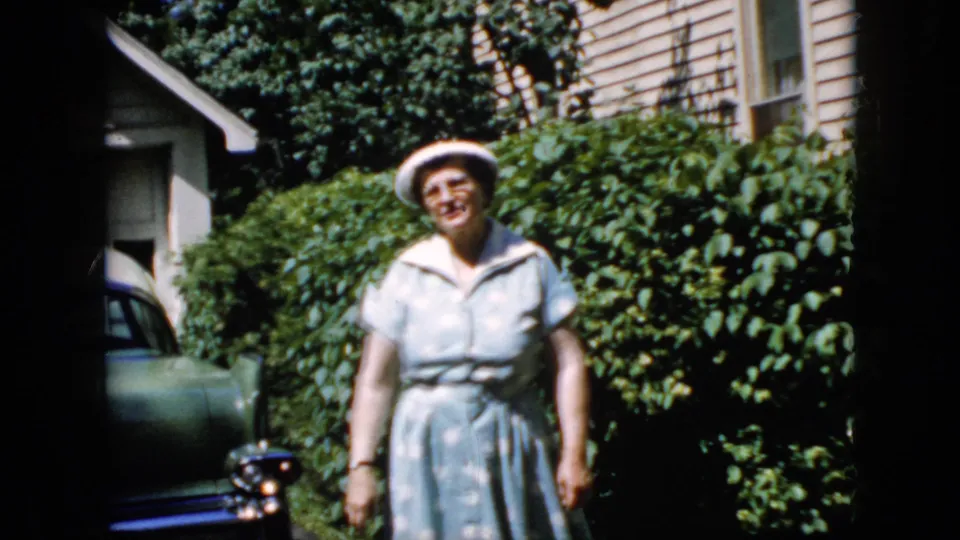Adult woman in a white hat and light blue polka-dot dress faces the camera in a sunlit California driveway, with a green car, hedges, and house siding behind her.