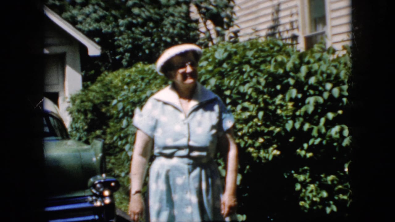 Woman in a white hat walks toward the camera beside hedges and a parked car.