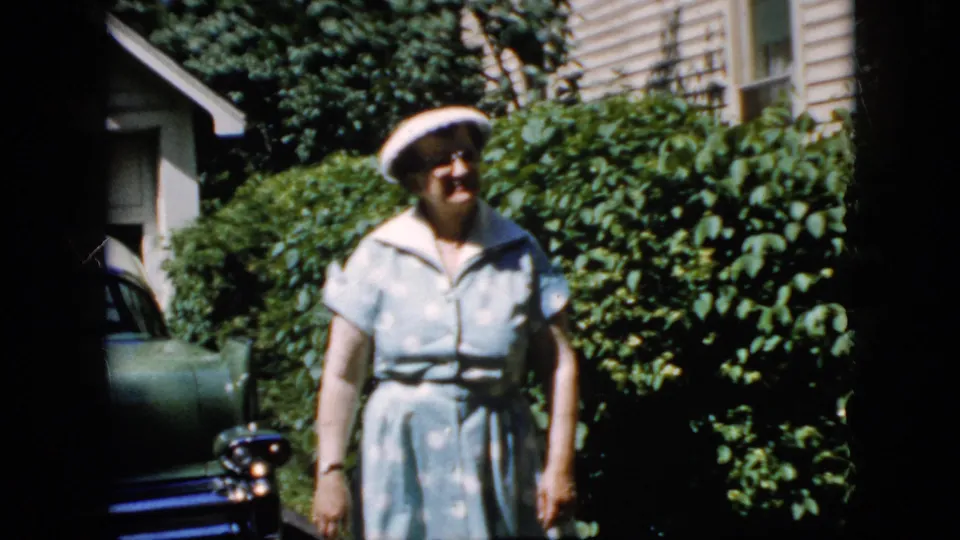 Adult woman in a white hat and blue dress walks toward the camera in a 1969 California driveway scene, with a green car partly visible at left and leafy hedges behind her.
