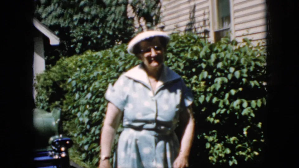 Adult woman in a white hat and light blue dress smiles as she walks closer through the sunlit driveway, with a parked green car and dense greenery framing the background.