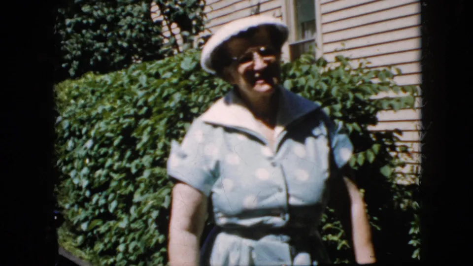 Closer 8mm frame of the adult woman smiling in her white hat and light blue polka-dot dress, with leafy hedges and residential siding visible in the California driveway setting.