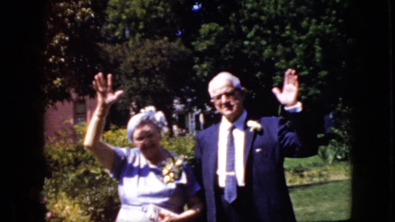 Senior couple standing in a sunny yard and waving toward the camera.