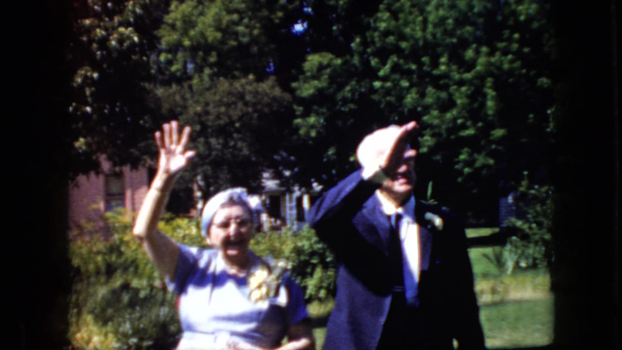 Senior couple continues waving, with the man's hand partly shading his face.