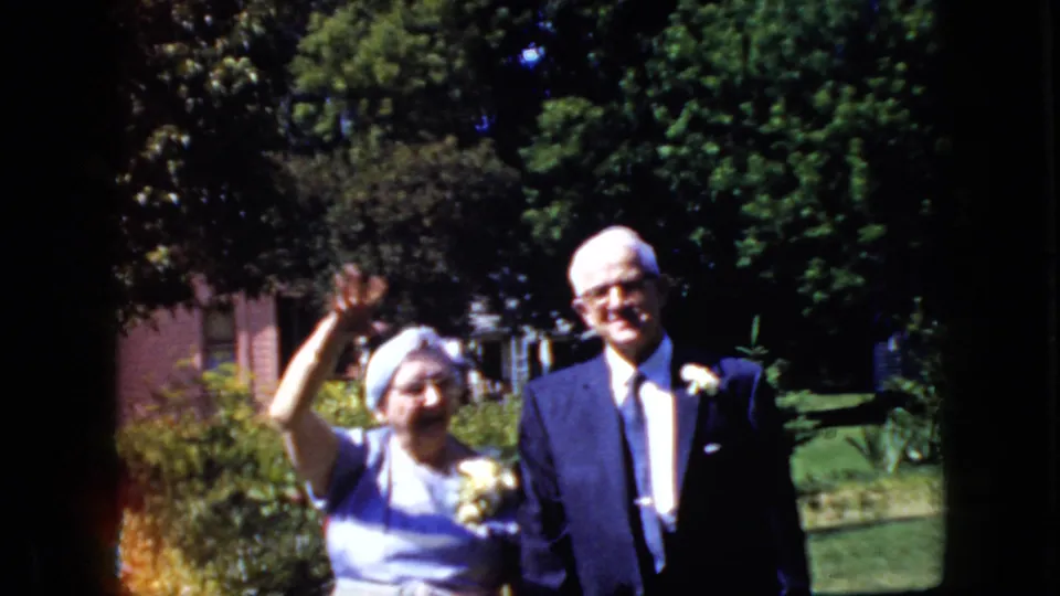 The woman in a light dress and white head covering waves beside the older man in a dark suit, with garden plants and a lawn behind them.
