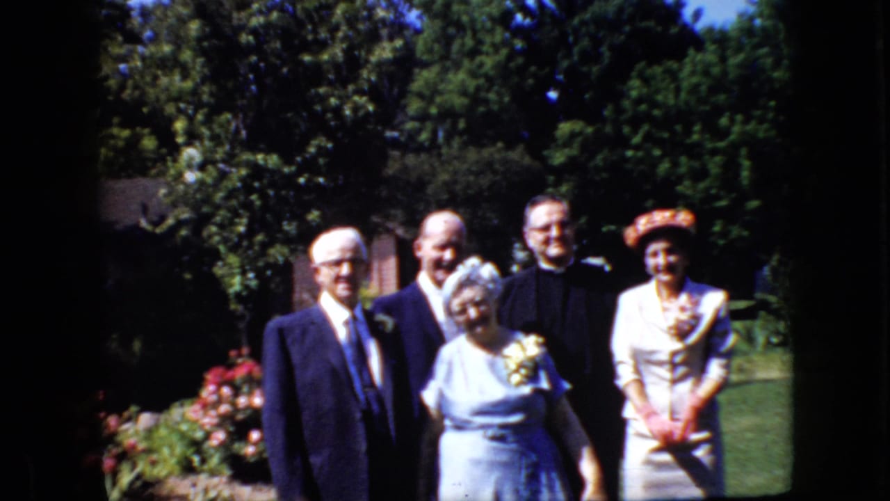 In supported 1969 California home-movie footage, five adults stand together in a garden for a formal group pose, with suits, clerical attire, a corsage, and a patterned hat visible.