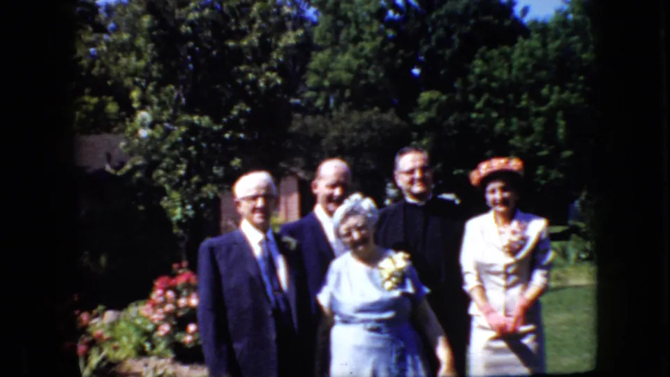 In supported 1969 California home-movie footage, five adults stand together in a garden for a formal group pose, with suits, clerical attire, a corsage, and a patterned hat visible.