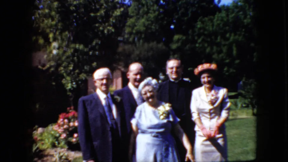 The group remains arranged in the sunlit garden, showing formal late-1960s clothing, a corsage, clerical dress, flowers, lawn, and trees in the background.