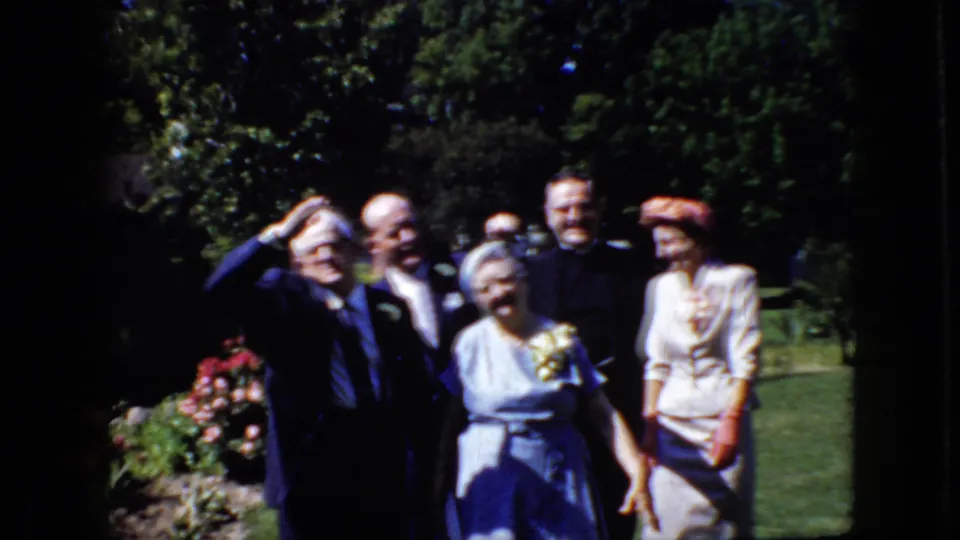 During the brief home-movie pose, one suited man raises his hand toward the camera while the others smile or look on in the formal garden setting.