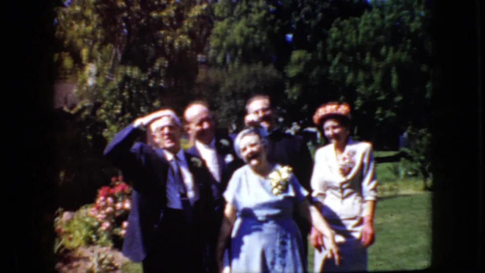The final frame shows the formal garden group relaxing from the pose, with faces visible, one man gesturing upward, and the women in blue and white standing near the front.