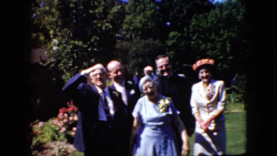 In this 1969 California home-movie frame, formally dressed adults stand in a garden with men in suits and women in dresses and hats facing the camera.