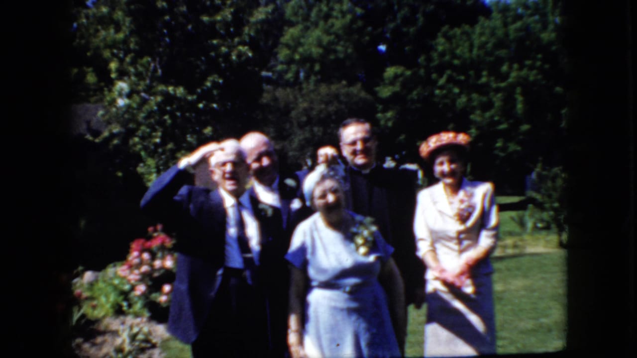Group of adults in suits and dresses stand closely together on a lawn beside flowering plants.