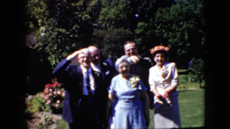 The group remains posed in the California garden setting, with formal suits, dresses, corsages, and dense greenery visible in the soft 8mm frame.