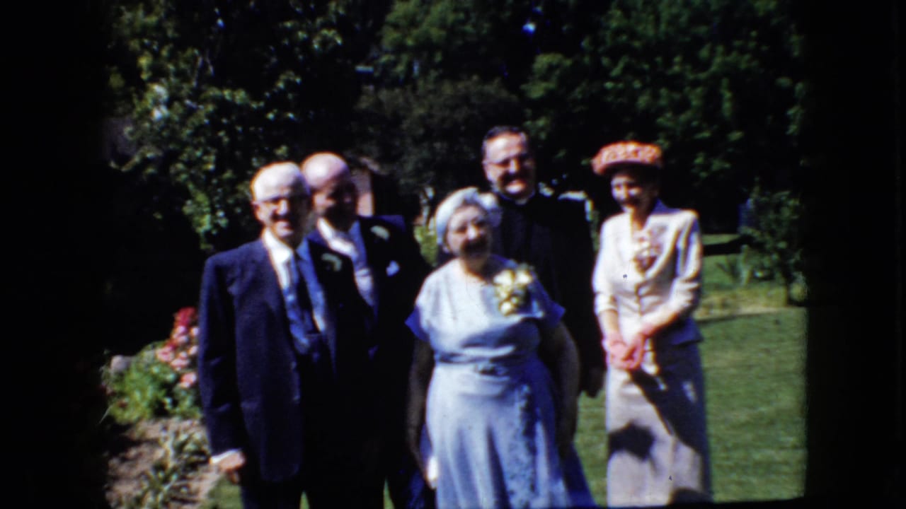 Older adults in suits and dresses pose together in a California garden setting from the supplied 1969 8mm home-movie clip.