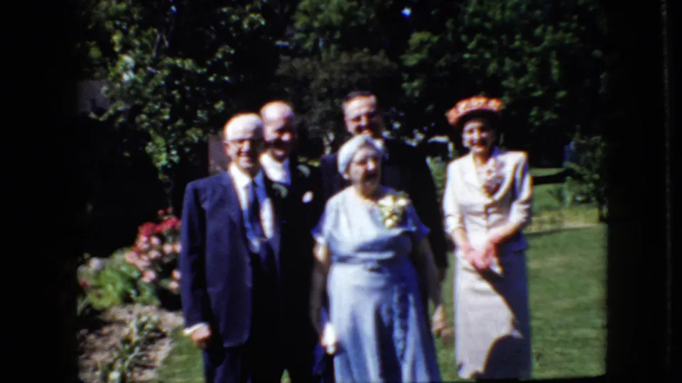 Five older adults in formal attire stand in bright sunlight on a garden lawn in the 1969 California home-movie scene.