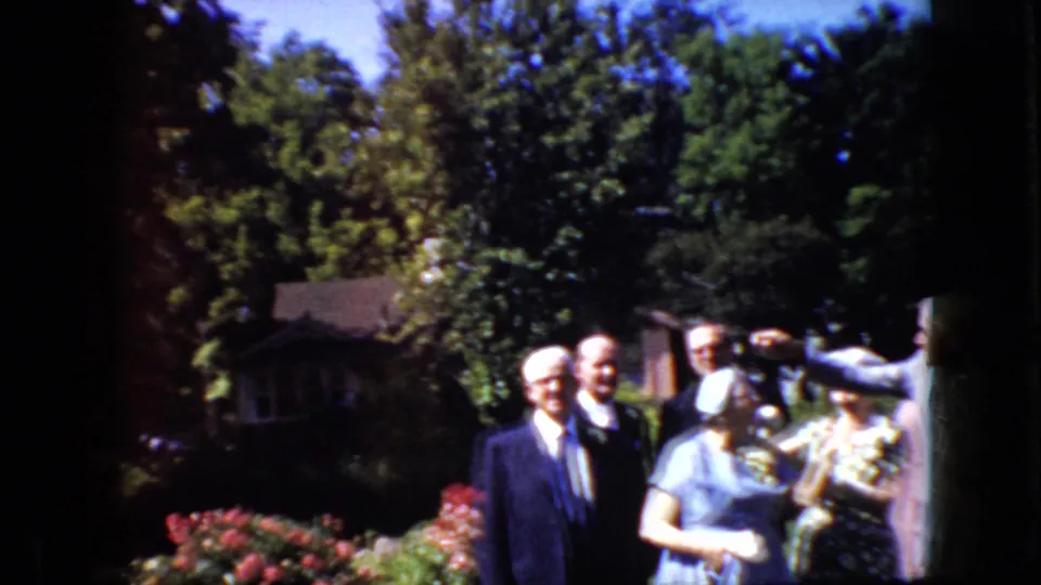 Older guests in formal clothing stand near flowers and trees as a partially visible person gestures from the right edge of the 1969 California garden footage.