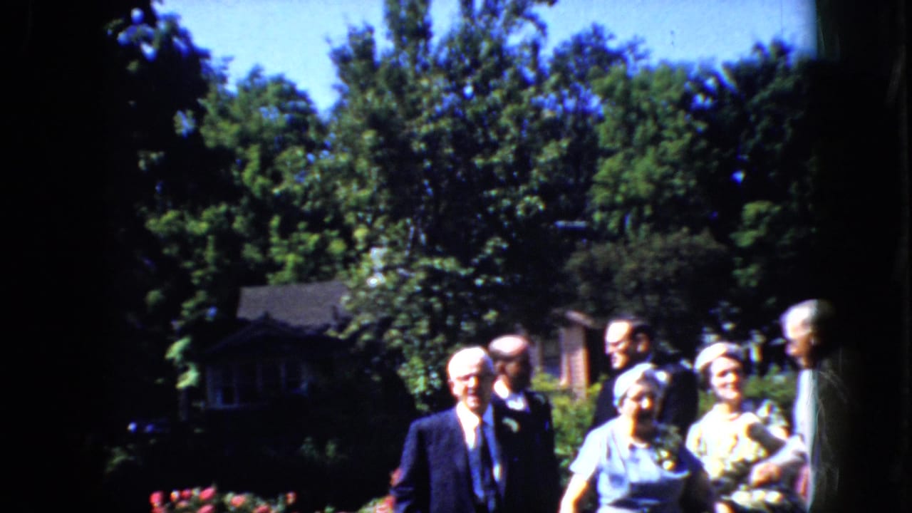 Older newlywed couple and guests stand outdoors in a sunny garden as the wedding group gathers.