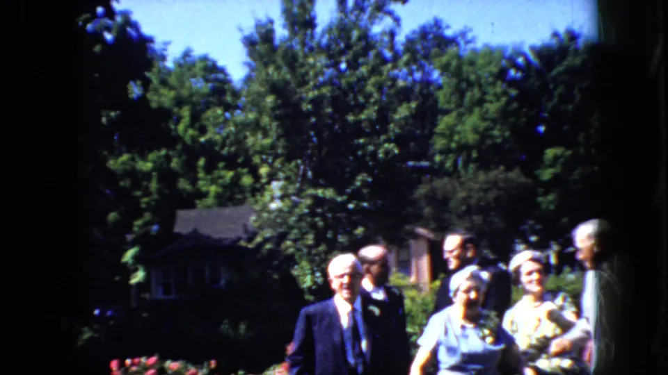 In 1969 California home-movie footage, an older bride in a light dress and hat stands near an older groom in a dark suit while guests gather in a leafy garden.