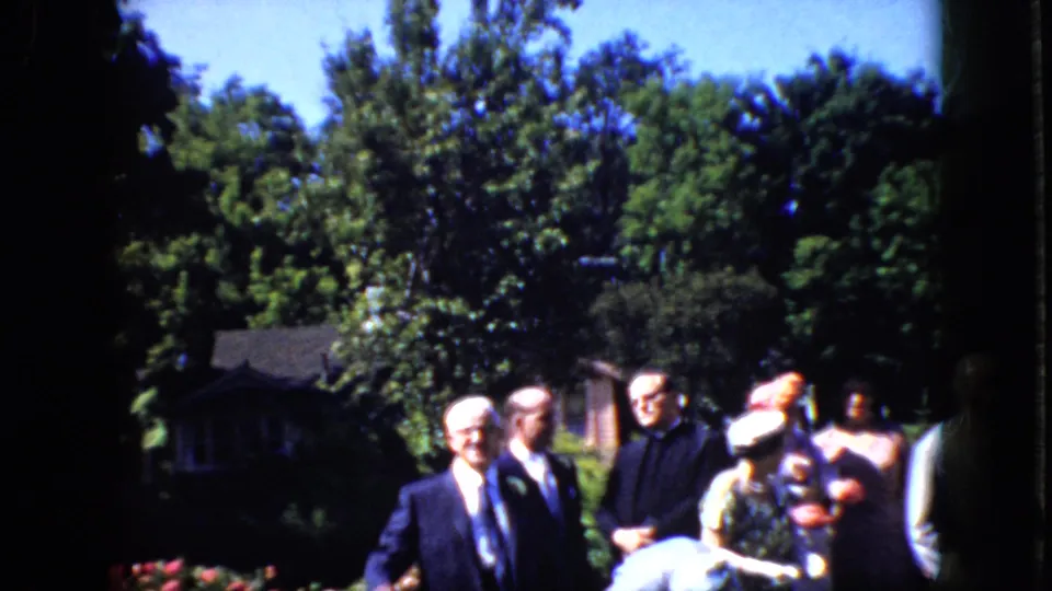 A handheld 8mm frame shows the older newlyweds partly surrounded by formally dressed guests during the rice-toss moment in a California garden setting.