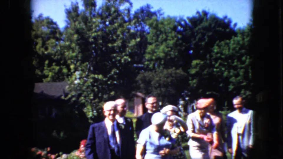 The older couple stands near the center of a 1969 California outdoor wedding scene as guests in suits, hats, and light dresses gather around them in bright sun.