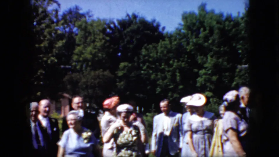 A small group of adults in late-1960s formal clothing moves through a California garden setting, with visible hats, patterned dress fabric, suits, and trees.