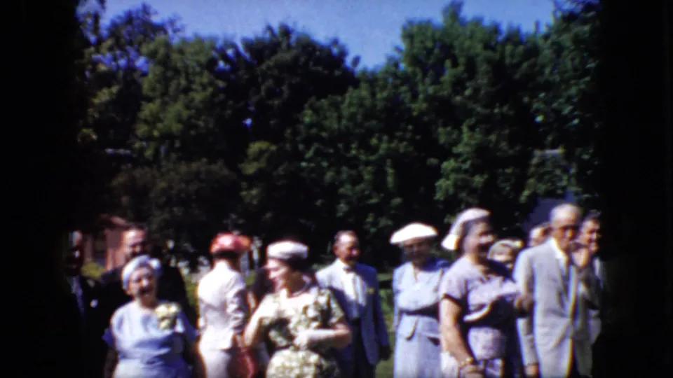The closing frame shows several formally dressed adults standing together outdoors in the supplied 1969 California home-movie footage, with greenery and bright daylight in the background.