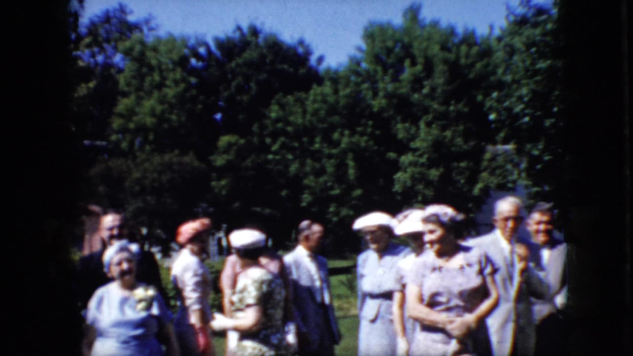 Older adults in formal clothing stand on a sunny lawn with trees behind them.