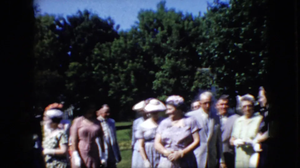 Older adults at a formal outdoor gathering in California appear in small clusters, with women in light dresses and hats and men in suits near trees.
