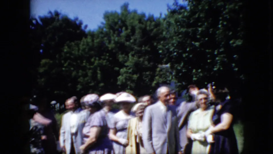 The home movie shows older guests in formal 1960s clothing gathered on a lawn, including a suited man and a woman in a pale dress near the front.