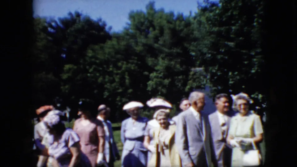A small group of older adults in suits, dresses, hats, and gloves stands outdoors in the 1969 California home-movie scene, some facing the camera and others turned aside.