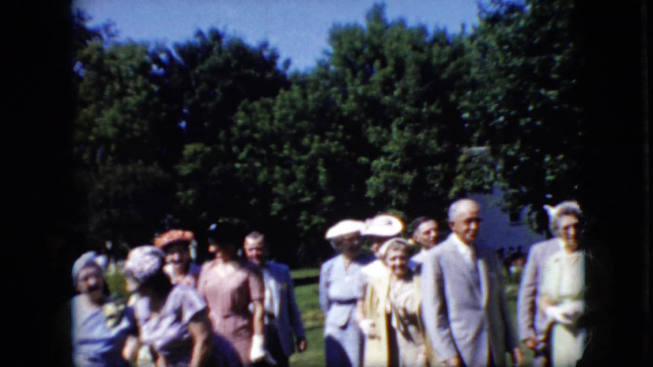 In California home-movie footage from 1969, older adults in suits, dresses, and hats gather on a sunlit lawn with leafy trees in the background.