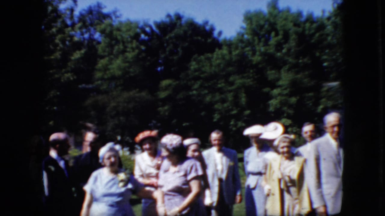 A group of formally dressed older adults stands in a park-like setting near trees.