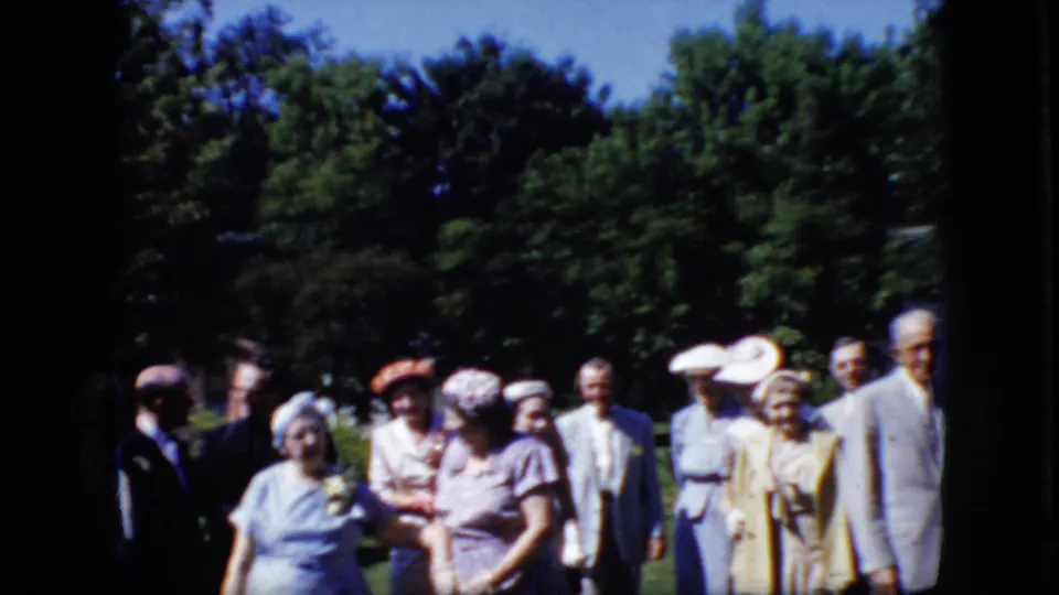Older adults wearing suits, dresses, hats, and pinned flowers appear gathered outdoors in 1969 California, with trees and lawn visible behind them.