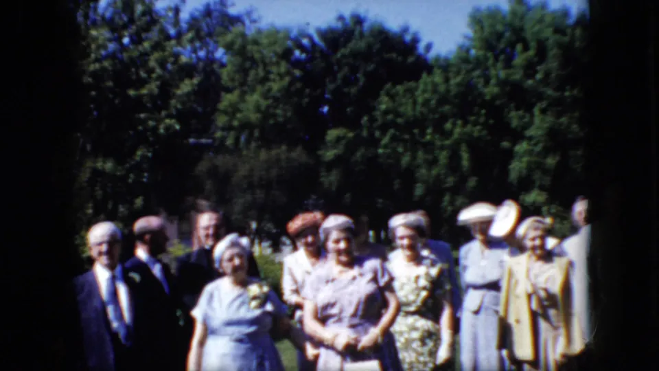 A home-movie frame shows older adults in Sunday-best clothing standing together on a lawn in California, with trees filling the background.
