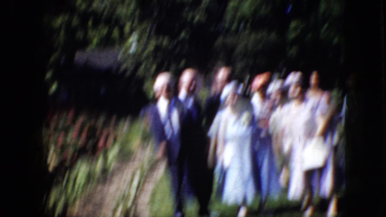 Blurred adults in formal clothing walking or standing outdoors near greenery.