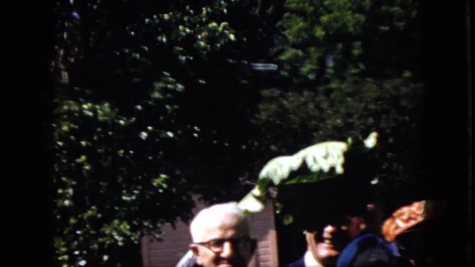 1969 California 8mm frame showing an older man with glasses among adults as a large palm frond is held up in front of dense greenery.
