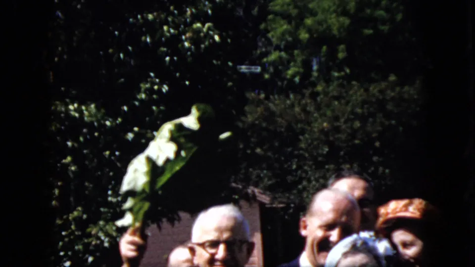 1969 California home-movie frame showing smiling adults in formal wear beside a raised palm frond against trees and garden foliage.