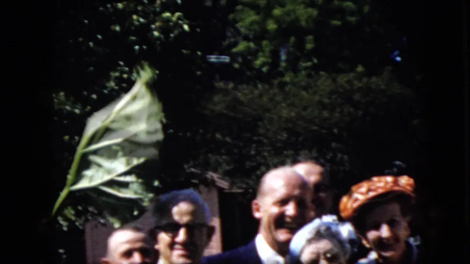 In this 1969 California 8mm home-movie frame, adults at an outdoor gathering smile toward the camera while greenery and a large nearby leaf fill the background.