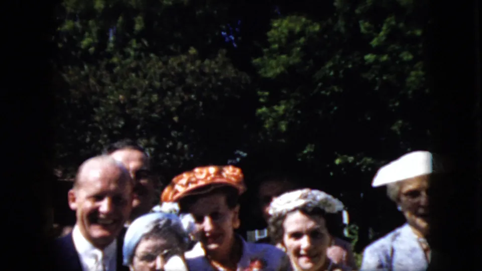 A 1969 California home-movie frame shows adults in suits, dresses, and hats clustered closely outdoors, smiling and leaning into the camera view.