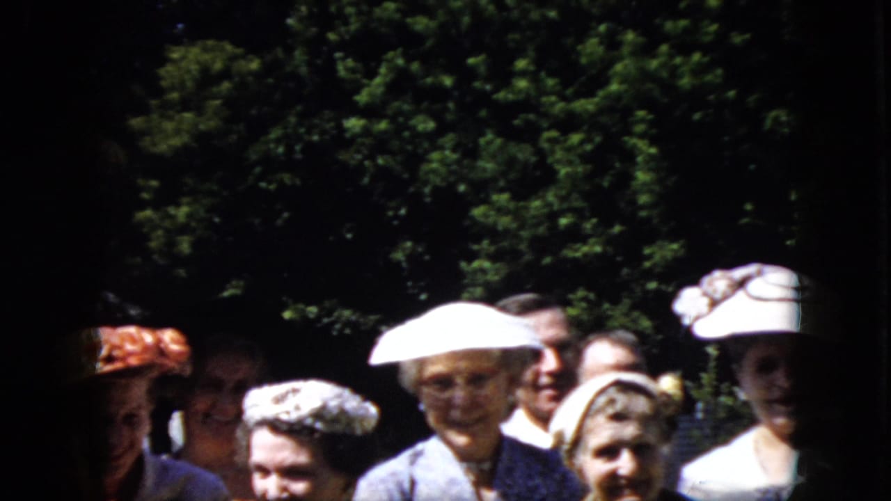 In 1969 California home-movie footage, older adults in white hats, suits, and dresses stand together in a shaded garden area.