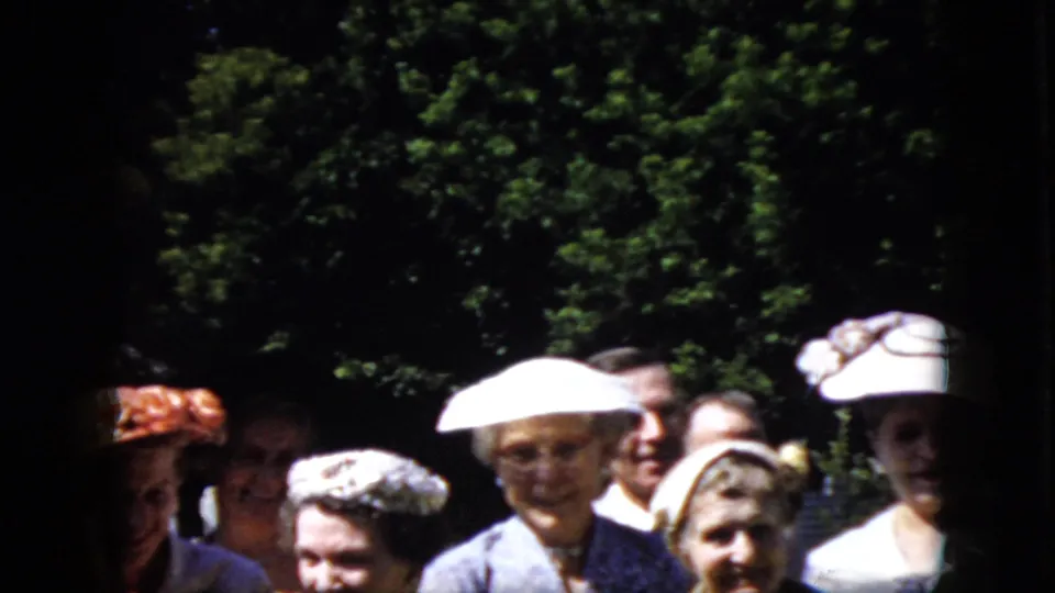 In 1969 California home-movie footage, older adults in white hats, suits, and dresses stand together in a shaded garden area.