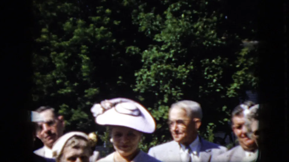 Older adults in formal clothing stand outdoors, with hats, suits, leafy trees, and part of a white building visible in the 1969 California home movie frame.