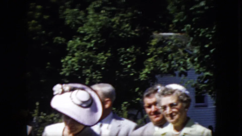 Older adults smile and stand together in a sunlit outdoor setting, with formal hats, light clothing, trees, and a white building visible in the background.