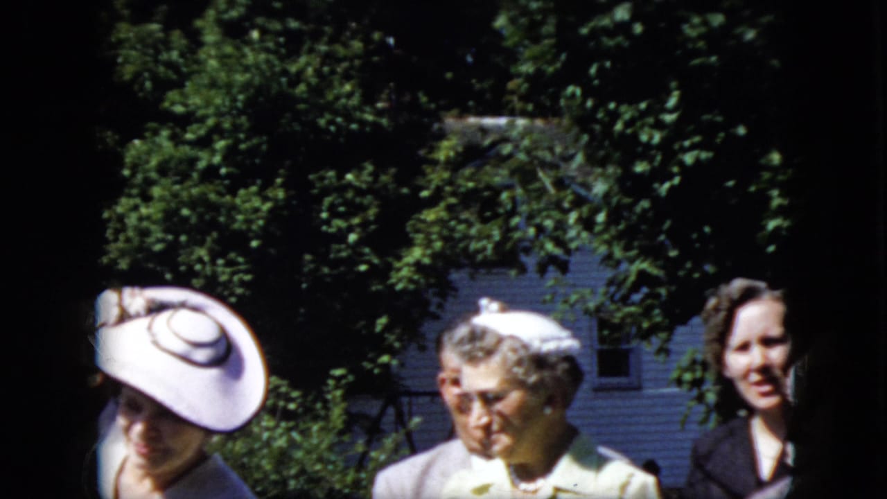 In 1969 California home-movie footage, women in formal hats and light-colored dresses stand outdoors in bright sun with leafy trees and a house behind them.