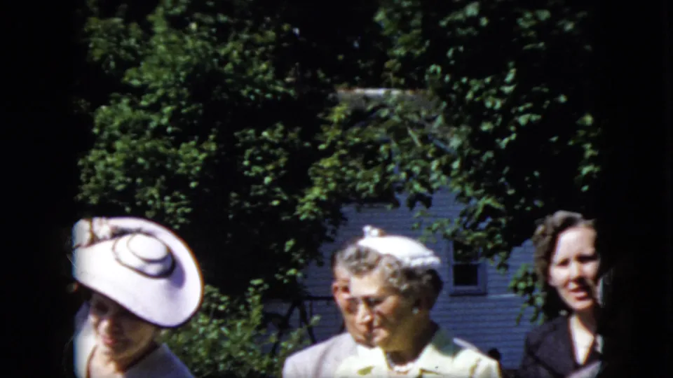 In 1969 California home-movie footage, women in formal hats and light-colored dresses stand outdoors in bright sun with leafy trees and a house behind them.