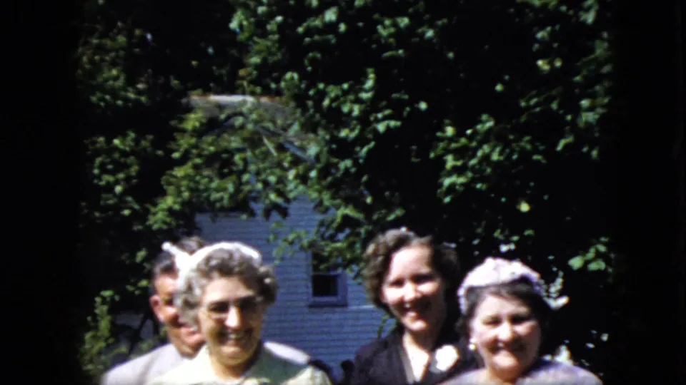 Adults at an outdoor California gathering in 1969 smile toward the home-movie camera, with women in formal dresses and hats framed by trees and a house.