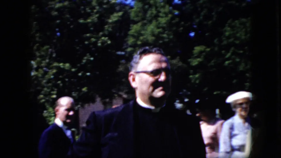 A man wearing clerical clothing appears in the foreground of 1969 California home-movie footage, with other formally dressed adults and trees in the background.