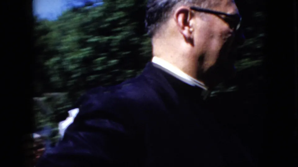 Close side view of a man in clerical clothing moving through a leafy outdoor setting in 1969 California home-movie footage, with motion blur from the handheld camera.
