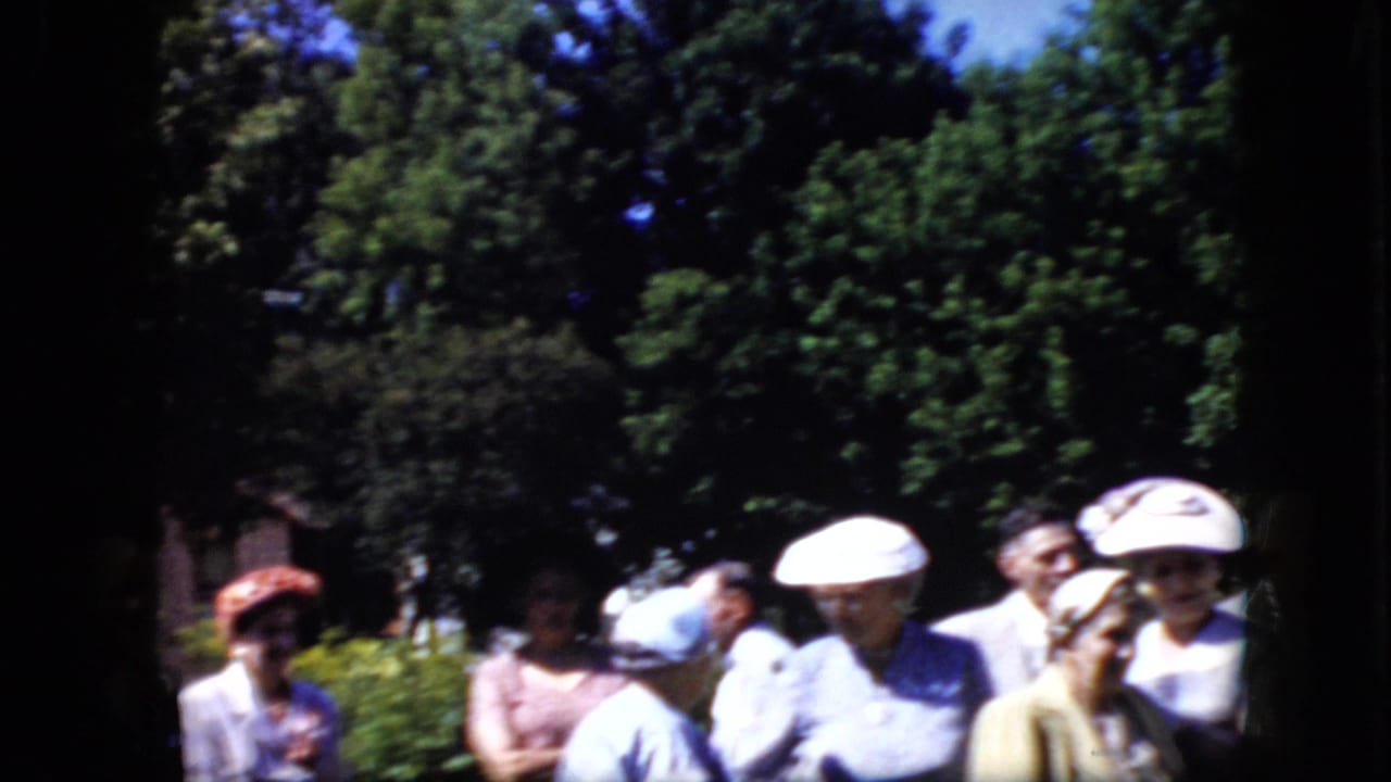 Adults in formal clothing and hats stand outdoors near trees and shrubs.